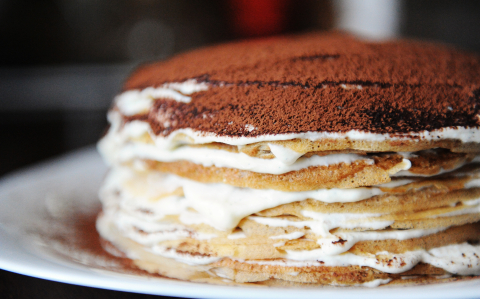 Freshly cooked crepe cake with tiramisu filling dusted with powdery cocoa. White plate, background of coffee house. Side view, close up